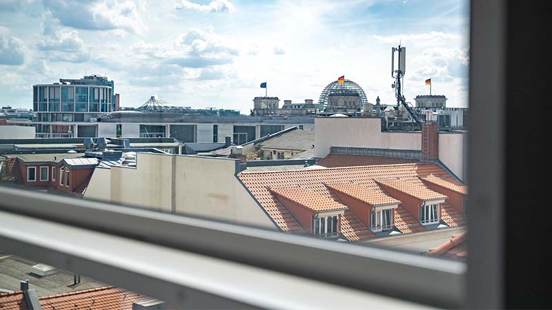 Blick vom 7. Stock auf Berliner Dachlandschaft mit Reichstagskuppel und Mobilfunkmast bei Tageslicht.